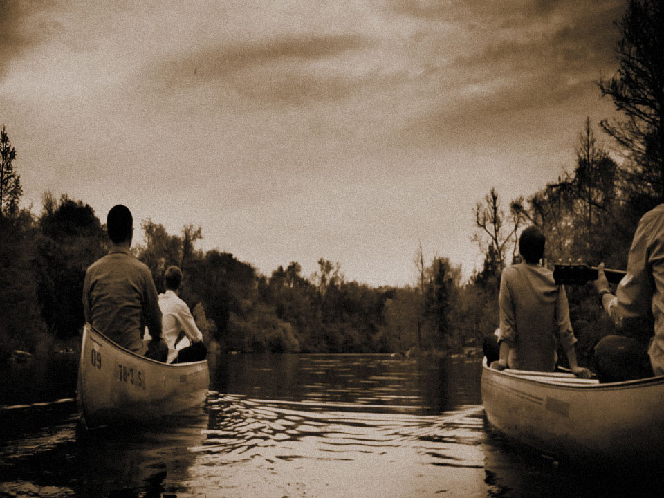 Two canoes drifting down a tree-lined river in a sepia-toned film still