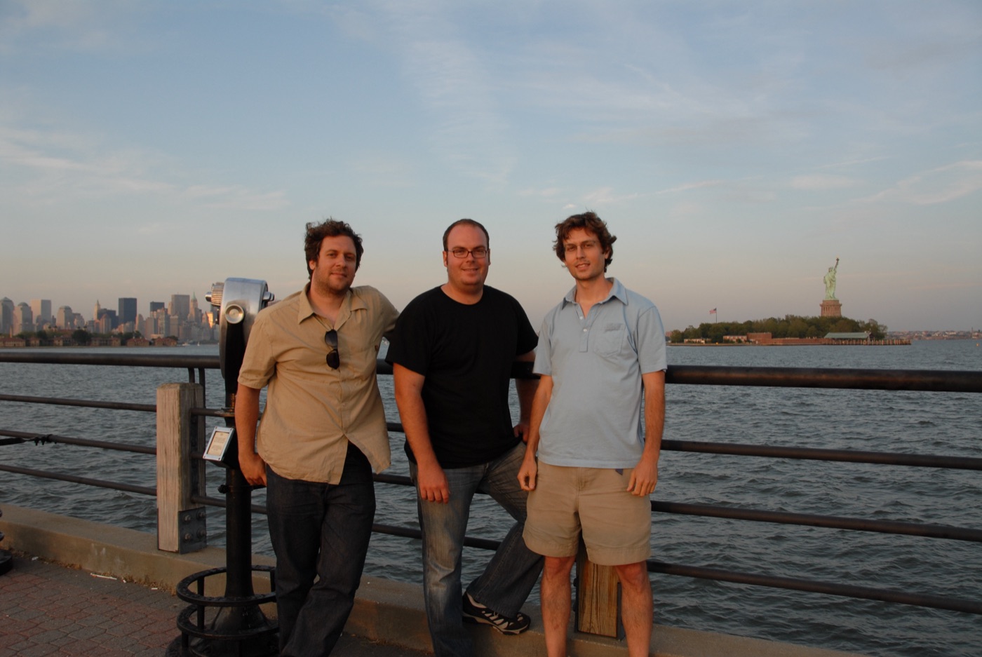Three crew members at a harbor railing with the Manhattan skyline and Statue of Liberty behind them