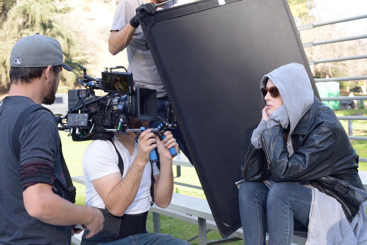 A camera operator lines up a shot on a hooded actress beside a large black flag outdoors
