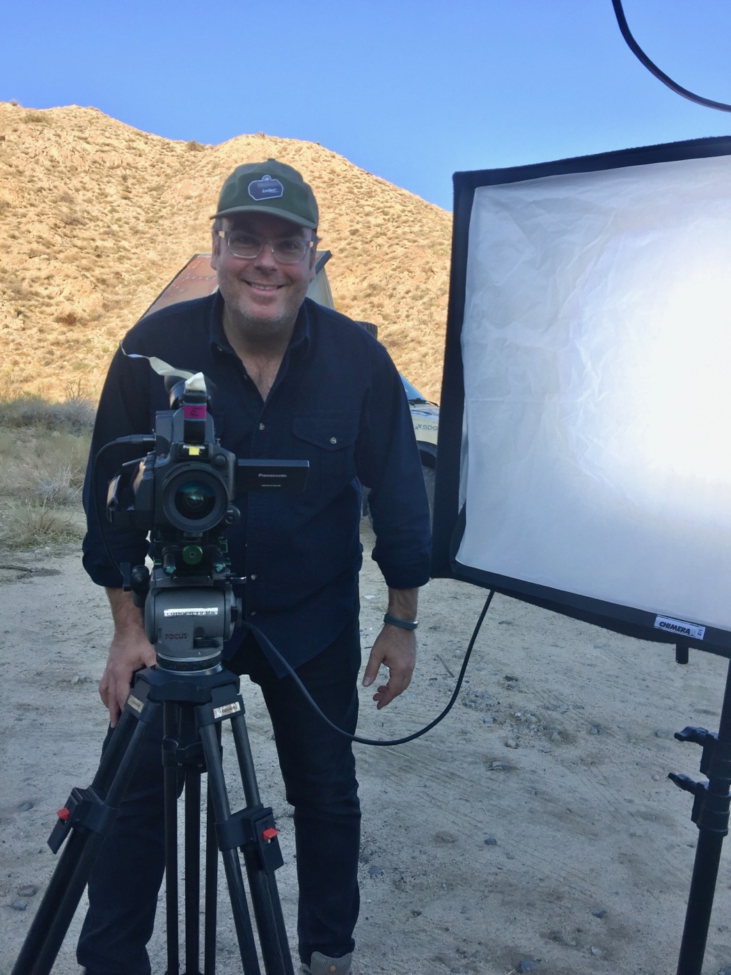 A cameraman in a Lodger cap behind a tripod-mounted camera and softbox on a desert location