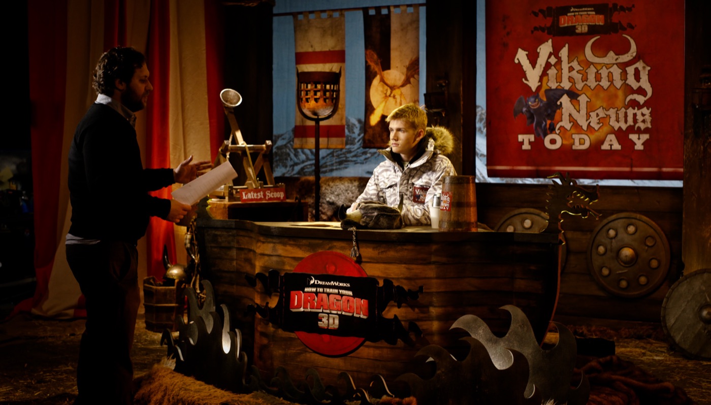 A director briefs the host at the anchor desk of the Viking News set for a How to Train Your Dragon promo