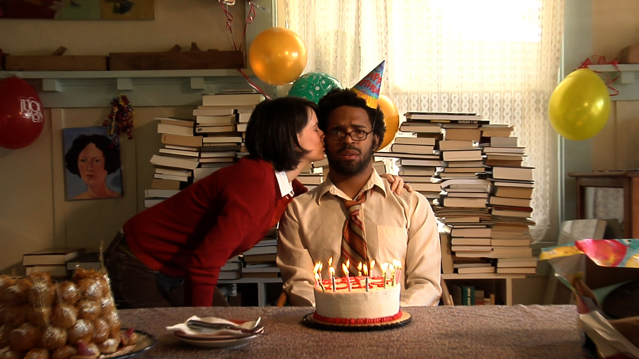 A woman kisses a glum man in a party hat behind a candle-covered birthday cake, stacks of books behind them