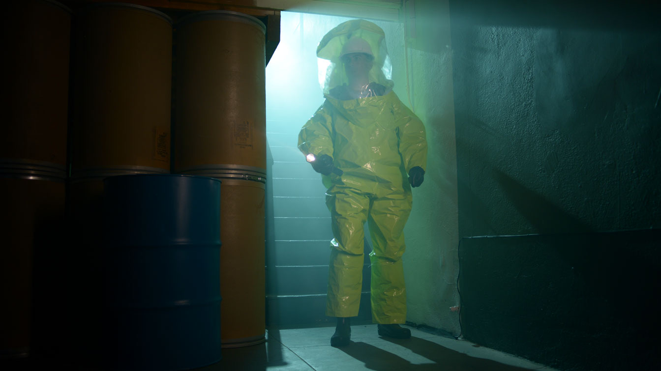 A figure in a yellow hazmat suit sweeps a flashlight through a fog-filled room stacked with barrels