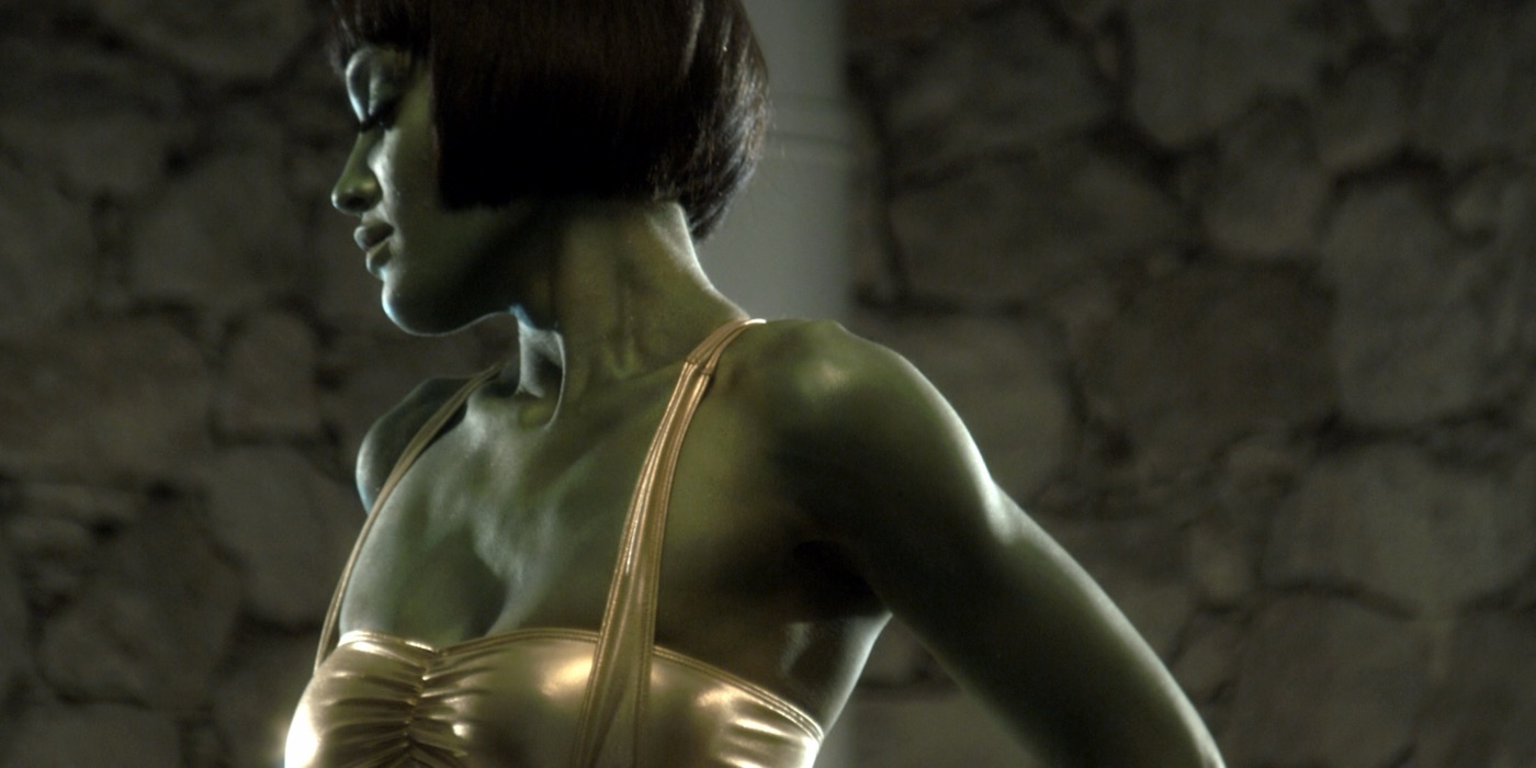 A performer in green body paint and a gold costume against a stone wall in a music video still