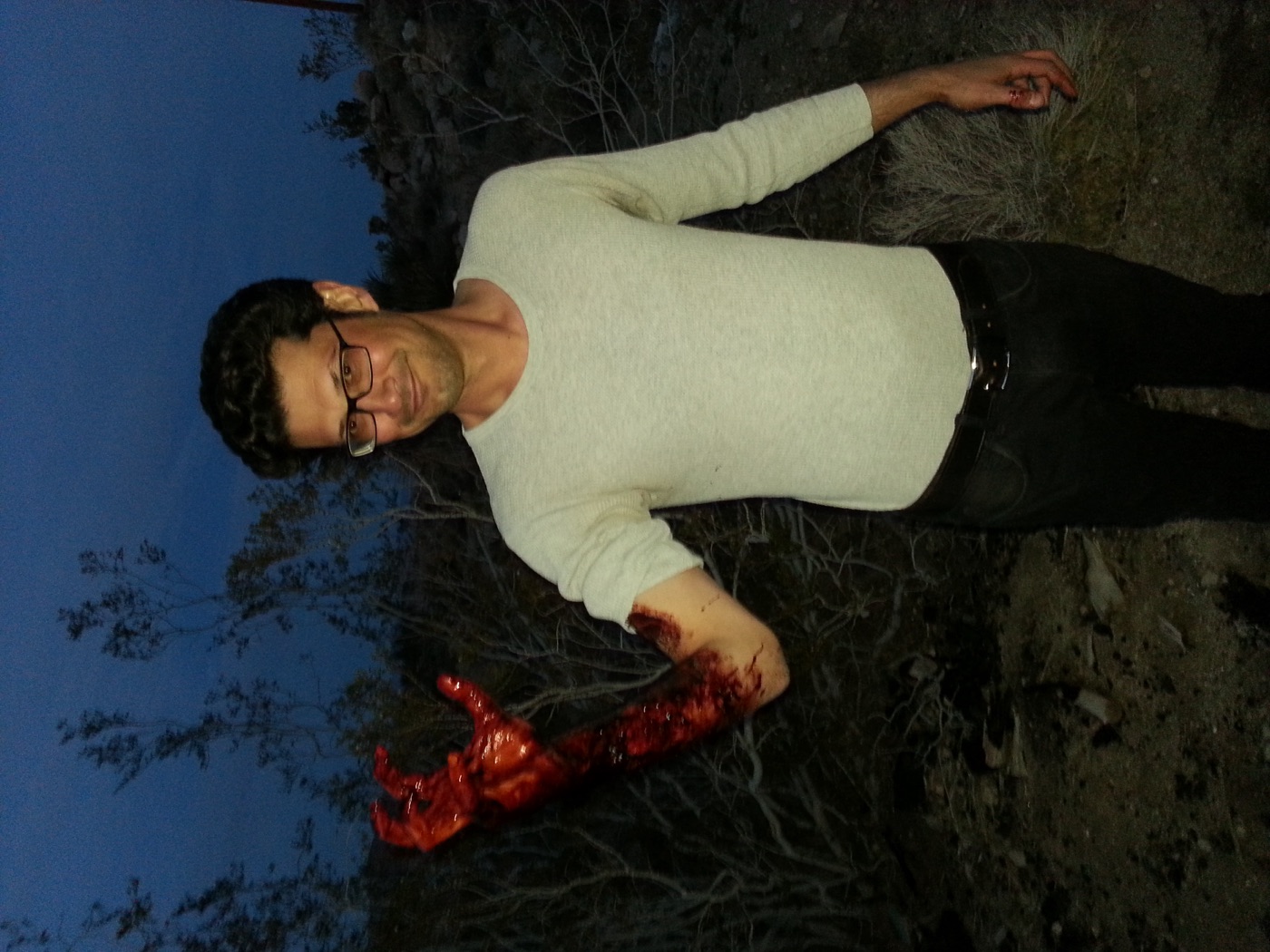 A crew member grins while holding up a bloody prosthetic arm effect at dusk on a desert location